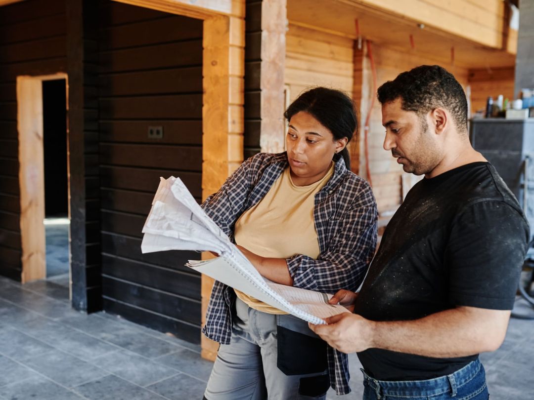 Zwei Personen halten einen Bauplan vor einer dunklen Holzfassade und besprechen ihn im Freien